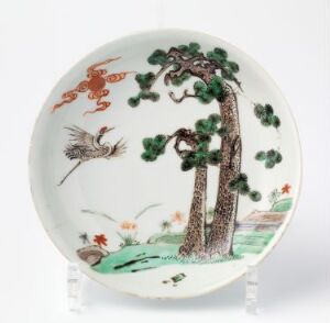  A decorative plate with a hand-painted scene of a large brown tree with green foliage on the right side, rolling green hills at the base, and a bird with orange and green plumage flying towards the tree, all on a white background.