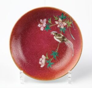  A round ceramic plate with a glossy burgundy surface featuring a detailed illustration of a bird on a branch with green leaves and pink blossoms, accented with gold speckles, displayed on a clear stand against a white background. Artistname and title are unknown.