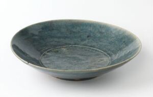  A ceramic bowl with a gradient of blues, ranging from pale turquoise at the center to a richer teal at the edges, with a semi-glossy finish against a plain background. Artistname and title are unknown.