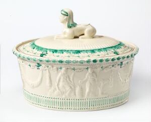  An antique cream-colored oval ceramic tureen with classical relief figures and green decorative accents, featuring a small figurine of a hound as the handle on the lid.