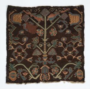  A square, vintage-looking textile with a central vertical stem and symmetrical patterns in muted colors of orange, green, blue, and cream on a chocolate brown background. The design features traditional, possibly historical, motifs and embroidered alphanumeric patches. Artist name and title unknown.