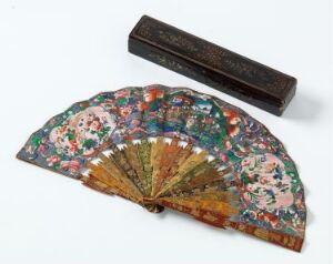  An opened, elaborately decorated folding fan with a colorful floral design on its upper half and a panoramic scene on the lower half, displayed next to its dark, possibly carved wooden case on a white surface.