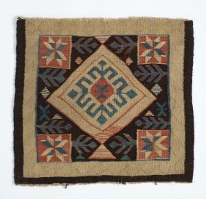  An image of a square traditional textile featuring a geometric quilt pattern with a tan border and a variety of earthy colors including blues, reds, oranges, and greens, showing star-like and diamond-shaped designs and frayed edges.