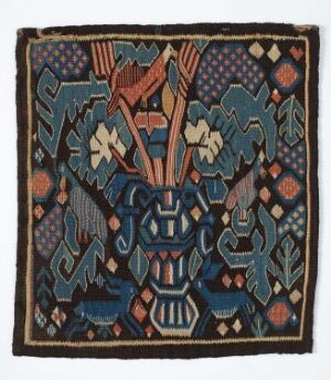  An intricately patterned textile with a central abstract mask-like figure surrounded by stylized birds and geometric designs in shades of blue, orange, beige, and brown, featuring a fringed border, indicating cultural or traditional artistry.