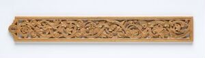  A long, narrow wooden panel with detailed, symmetrical carvings, possibly of floral or foliate designs, set against a white background. The craftsmanship is intricate, with the carvings giving a three-dimensional effect to the warm light brown material.