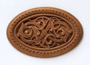  An intricately carved, oval-shaped reddish-brown object with a textured border and a complex central pattern resembling Celtic knots, set against a light-colored background.