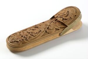  An intricately carved wooden object with a floral pattern and a honey-brown finish on a reflective white surface. The object resembles a traditional hairbrush and reflects light off its smooth and detailed surface.