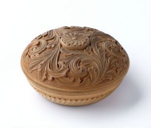  A small, detailed ceramic box with a domed, intricately carved lid, displaying floral and swirl patterns in a warm earthy brown color, against a white background. Artist name and title are unknown.