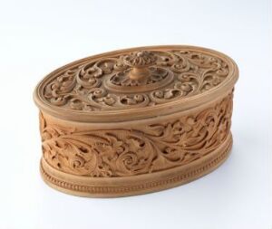  An intricately carved oval wooden box with a detailed floral and scrollwork pattern, set against a plain white background. The warm brown tones of the wood highlight the craftsmanship and artistry of the carving.