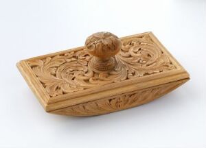  A carved, rectangular wooden box with intricate floral designs and a round handle on the lid, set against a plain white background. Artist and title are unknown.