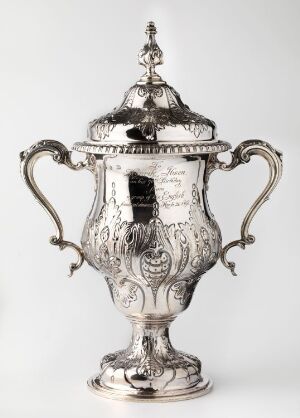  An ornate silver trophy cup with detailed etchings and two curved handles on a round pedestal, complete with a matching lid topped with a decorative finial.