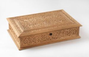  An intricately carved light brown wooden box with a hinged lid on a neutral background, featuring detailed floral and geometrical patterns and a small central lock.