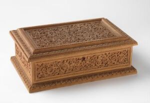  An intricately carved wooden box with a closed hinged lid, showcasing elaborate lace-like floral and geometrical patterns on a reddish-brown wood, photographed against a light, neutral background. Artistname and title unknown.