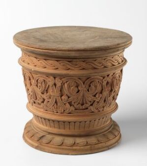  An intricately carved wooden side table or stand with a flat, circular top and a richly detailed cylindrical body featuring stylized floral motifs. The object exudes natural wooden color tones ranging from light beige to deep brown. Artist name and title are unknown.