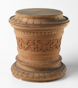  A cylindrical, intricately carved wooden pedestal with a series of decorative bands including a detailed floral motif in the center, set against a plain white background. Artistname and title are unknown.