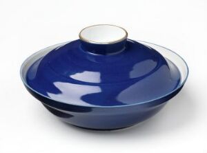 
 A glossy cobalt blue ceramic bowl with a lid, featuring a contrasting light-colored rim and knob, set against a neutral gray background. Artist name and title are unknown.