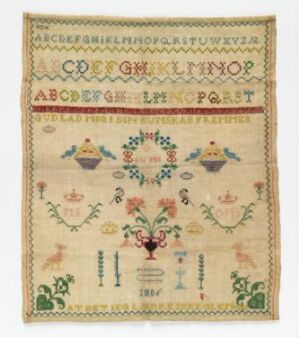  A traditional hand-stitched sampler featuring faded alphabets, numbers, and symmetrical floral and avian motifs on a cream-colored background, reflecting historical needlework practices.