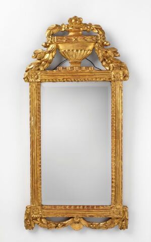  An antique vertical mirror with an ornately carved, gold-colored frame featuring classical decorations, including an urn at the top and intricate scrollwork.