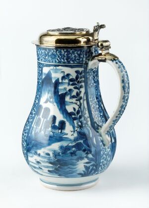  A traditional ceramic jug with a blue and white pattern, featuring a metallic lid and a smooth, curved handle, set against a plain background.