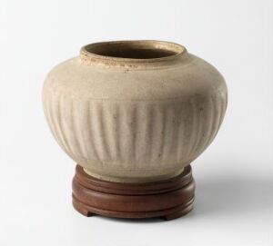  A round, grooved ceramic vase with a muted sand-colored body and darker brown top, placed on a dark brown wooden stand against an off-white background. Artist and title are unknown.