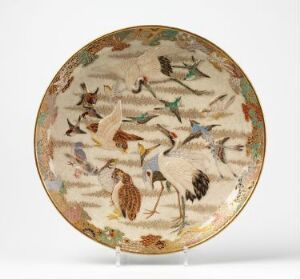  A round, decorative ceramic plate on a display stand, intricately painted with a complex scene of various bird species in natural poses, featuring a pale, creamy background with a border that may have gold accents, and a rich palette of browns, tans, blues, greens, and reds in the depiction of the birds and foliage.