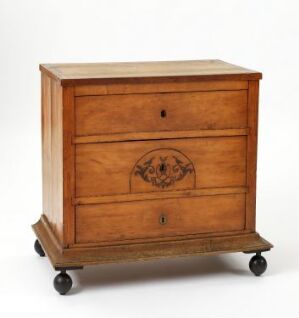  An antique wooden nightstand with a narrow top drawer, a larger bottom drawer featuring a circular carved medallion, on small spherical wheels, against a plain off-white background.