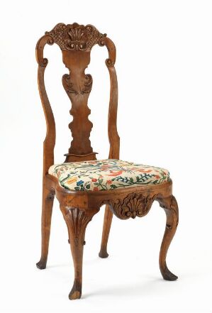  An antique-style wooden chair with a high, ornately carved back and a floral upholstered cushioned seat. It features Queen Anne style cabriole legs and a decorative 'X'-shaped stretcher, set against a plain background.