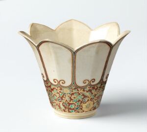  A decorative vase with a scalloped edge and intricate floral designs in red, blue, green, and yellow on a creamy off-white background. The vase resembles a blooming flower and exhibits a traditional and handcrafted aesthetic.