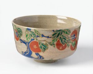  A handcrafted ceramic bowl with a cream or light beige background, adorned with a painted design of dark blue branches, green leaves, and round orange fruits with red symbols. The unpainted rim and bottom show the rustic brown of the pottery material.