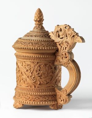  An ornate light brown ceramic jug with intricate carvings, featuring a large, openwork handle and a domed lid with a pointed top, against an off-white background. Artistname and title are unknown.