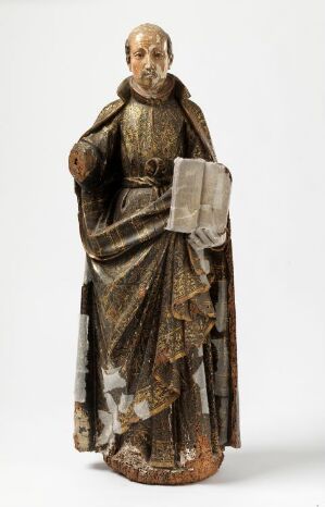  A carved wooden figurine of a bearded male saint in draped robes, with faded gold accents and an open book in one hand, standing on a round base with an aged and worn appearance.