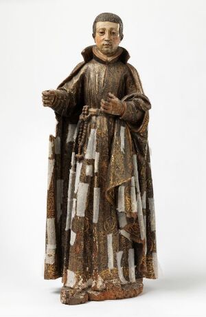  A figurine of a Franciscan friar or monk standing upright in traditional brown habit with a white cord and suspended beads around the waist, the right hand raised in a gesture, the left hand holding a book, against a neutral off-white background.