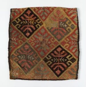  An intricately patterned square textile piece with fringed edges, showcasing a rich color scheme of deep reds, burnt oranges, greens, and golds arranged in diamond shapes with traditional designs.