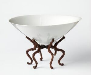  A unique white bowl with a thin rim and reflective surface is supported by a stand made of intertwined dark brown branches or roots, contrasting the bowl's modern look with a natural, rustic base.