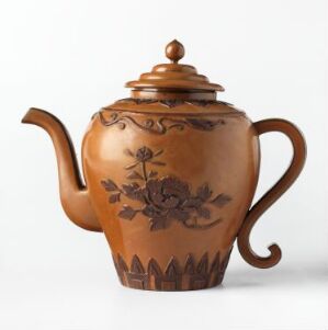  A glossy brown ceramic teapot with a decorative floral pattern and a pointed fence-like design around the base, set against a plain light background. Artist name and title are unknown.