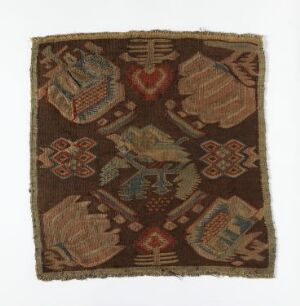  A fragment of an old textile, possibly a historical rug or tapestry, displaying symmetrical patterns with stylized bird-like figures and geometric floral designs in a color palette of earthy brown, burgundy, green, beige, and blue. The edges appear worn and frayed. Artist name and title are unknown.