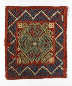  A detailed woven textile with a rich burgundy background and symmetrical pattern featuring blues, greens, and light beige. The design includes a central diamond-shaped medallion, smaller corner motifs, and intricate borders.