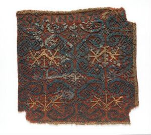  An ancient textile fragment with a complex geometric pattern in shades of dark brown, red, orange, teal, and pale yellow. The design features symmetrical star-like central figures surrounded by interlocking abstract floral motifs.