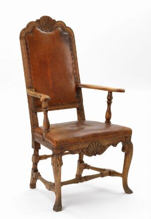  An image of an antique wooden chair with a tall, scalloped top edge and floral carvings on the backrest, a solid wood seat, turned spindles supporting the arms, and ornately turned front legs against a neutral background.