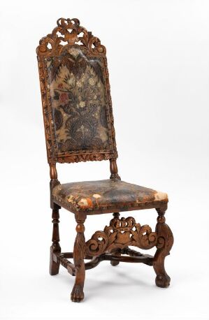  An antique chair with a tall, elaborately carved wooden backrest and matching legs, upholstered with a floral-patterned fabric in warm tones. The chair is set against a neutral background with a soft shadow to one side.