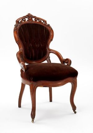  An antique mahogany armchair with deep brown velvet upholstery, embellished with a carved decorative crest on the back, curved armrests, and small casters on the front legs.