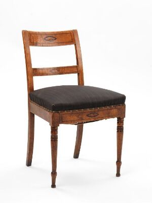  A traditional wooden chair with turned front legs, a padded black upholstered seat, and a decorative oval inset in the backrest, set against a light gray background. The artist name and title are unknown.