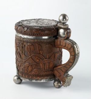  A decorative, cylindrical mug with three spherical feet and a handle, adorned with relief carvings of figures and foliage in dark and light brown tones, possibly a Renaissance-era artifact or a reproduction.