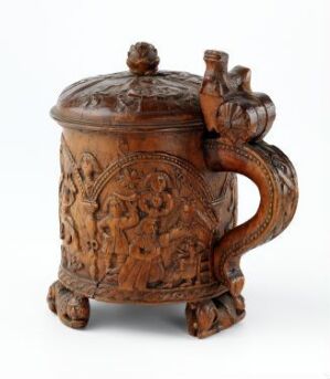  A beautifully carved birch wood drinking jug, titled "Drikkekanne," by an unidentified artist, featuring detailed relief scenes and figures on its cylindrical body, with a matching carved lid and an elegantly curved handle, all resting on three small spherical feet. The jug exudes a warm cinnamon brown hue, highlighting the intricate craftsmanship typical of traditional Scandinavian design.