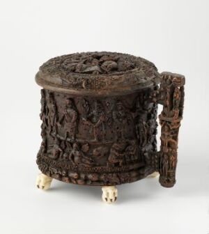  An intricately carved cylindrical wooden container or stool with three legs shaped like elephant feet, adorned with detailed bas-relief carvings of human and animal figures covering its entire surface, set against a white background.