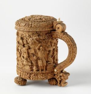  An ornately carved drinking vessel, titled "Drikkekanne" by an unidentified artist, made from masurbjørk—a figured birch wood—with relief scenes of human figures and details of ivory, set against a neutral background.