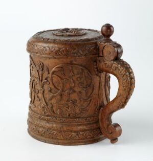  A wooden beer mug with a lid, intricately carved with natural and heraldic patterns in a monochromatic brown palette, displaying a traditional or antique style.