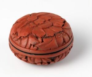  A small, round, reddish-brown carved wooden box with a pattern of overlapping leaves or petals covering its surface, showcasing detailed craftsmanship and a warm, inviting color palette. Artist name and title of the piece are unknown.