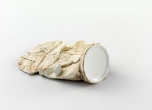  "Street-ware" by Kjell Rylander is an artistic ceramic piece showing a porcelain object with a rough, aged paper-like exterior in off-white and cream colors, contrasted by a section of smooth white porcelain.