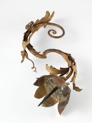  A copper-toned metallic sculpture resembling a twisting plant or vine with detailed leaf shapes, exhibiting a warm golden-brown color and a patina that gives it an aged look, displayed against an off-white background. Artist name and title are unknown.